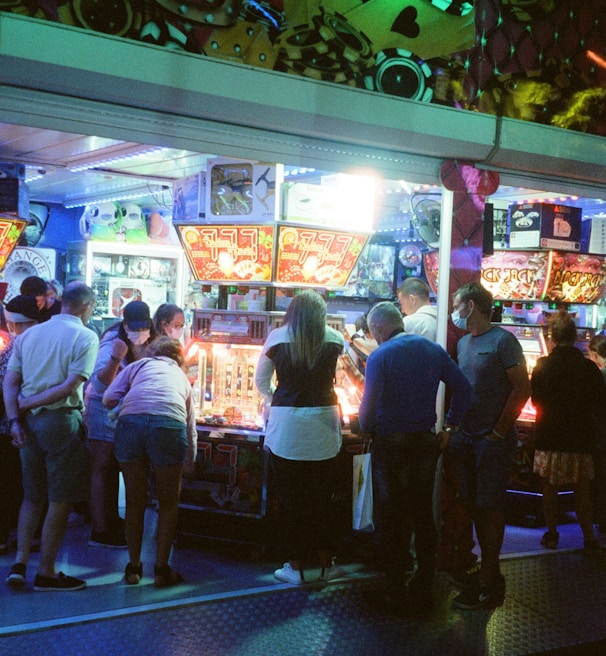 A group of people standing in front of an illuminated amusement arcade, engaging with various gaming machines. The environment is bustling with activity, and colorful lights create a vibrant atmosphere. The ceiling is decorated with large, playful images of dice and other game-related graphics, enhancing the lively mood.