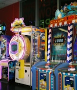 A row of brightly colored arcade game machines is arranged in a line against the wall. Each machine features vibrant lights and designs, with themes ranging from claw games to game show-inspired setups. The area has a playful and inviting atmosphere, suitable for entertainment or amusement centers.