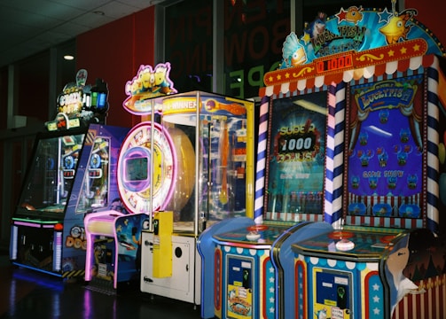 A row of brightly colored arcade game machines is arranged in a line against the wall. Each machine features vibrant lights and designs, with themes ranging from claw games to game show-inspired setups. The area has a playful and inviting atmosphere, suitable for entertainment or amusement centers.