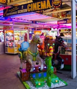 A vibrant amusement arcade featuring various games and bright neon signs. Mechanical horse rides are in the foreground, surrounded by claw machines with plush toys. The entrance is adorned with signs for a cinema and arcade, creating a lively and colorful atmosphere.