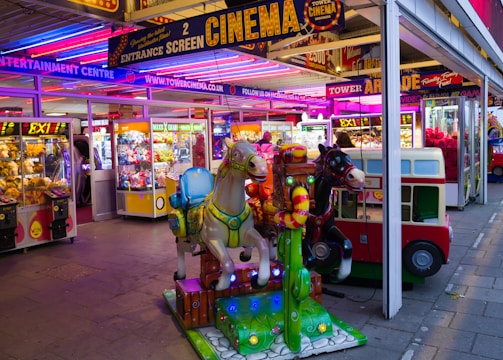 A vibrant amusement arcade featuring various games and bright neon signs. Mechanical horse rides are in the foreground, surrounded by claw machines with plush toys. The entrance is adorned with signs for a cinema and arcade, creating a lively and colorful atmosphere.